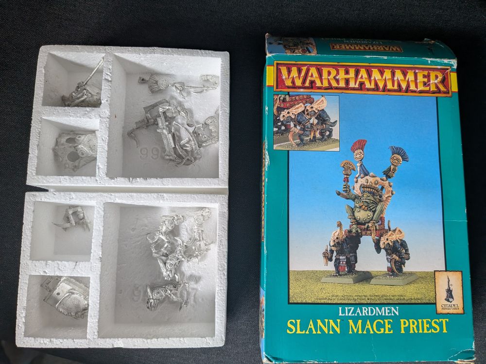 A photo of the box and contents for an unassembled classic Lizardman Slann Mage Priest model (big green frog guy being carried on a palanquin by blue lizardmen).