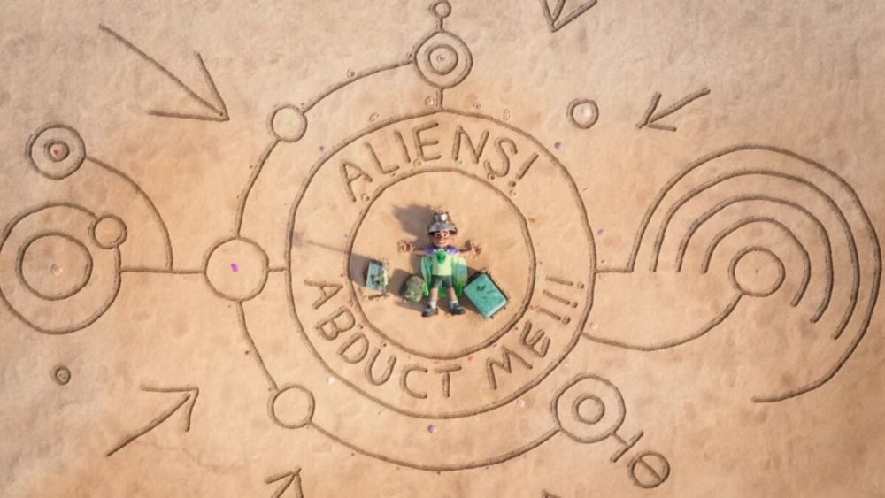 A still from "Elio". The main character lies on a beach with a sign saying "Aliens! Abduct me!!!"