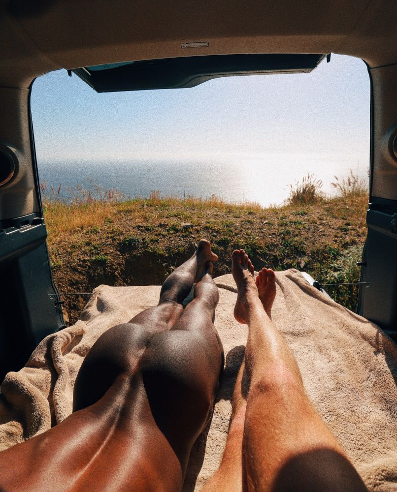 My legs and a guy’s backside lying down in the back of an SUV with the back open to the ocean view on a cliffside in Big Sur