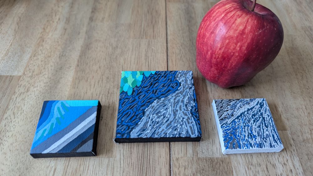 Three tiny canvases sit by a regular sized apple. The apple is used to show the small size of the three canvases.