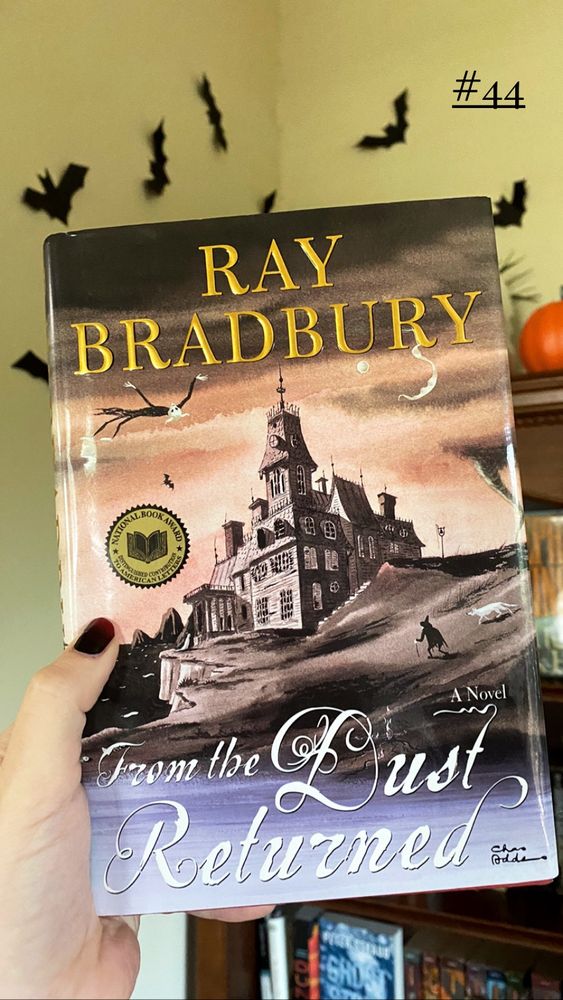 The cover of From the Dust Returned, which features a drawn picture of an old creepy mansion with a creature flying in the sky by the yellow name Ray Bradbury. I am holding it in my hand and in the background are black bats on my living room wall.