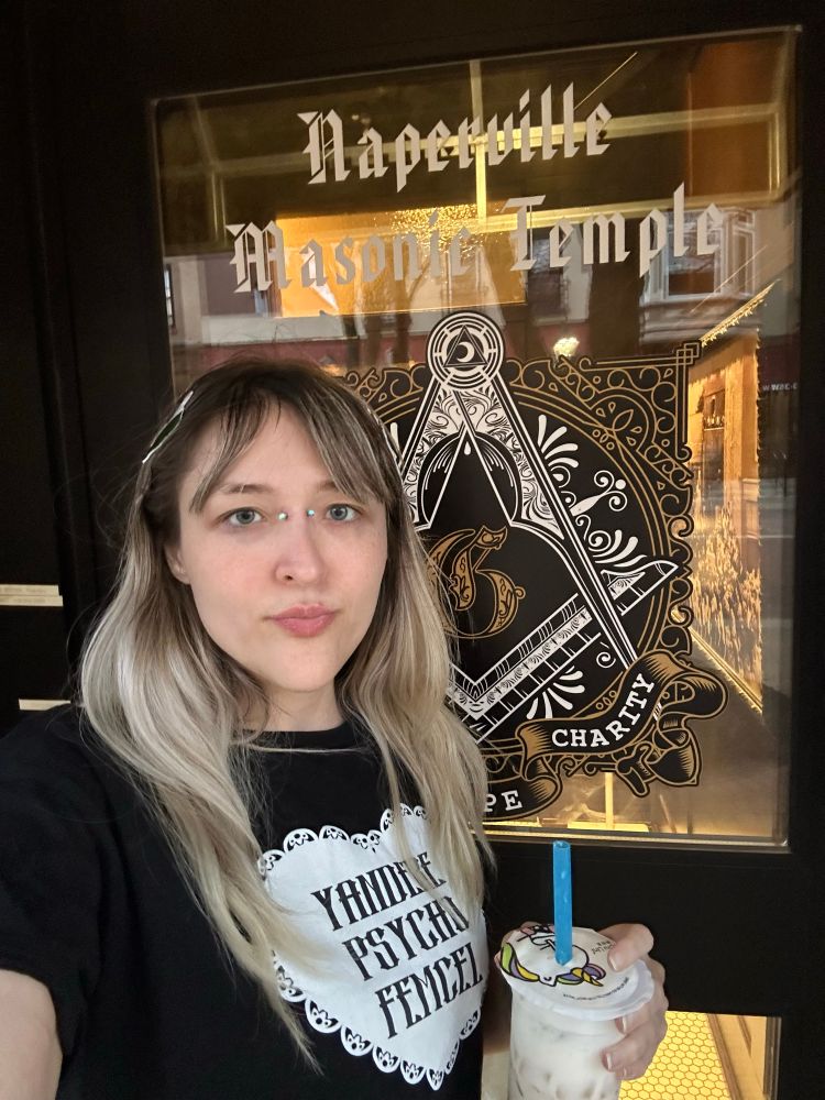 girl with piercing holding boba, wearing a shirt that says “yandere psycho femcel”, standing beside a door called “naperville masonic temple”