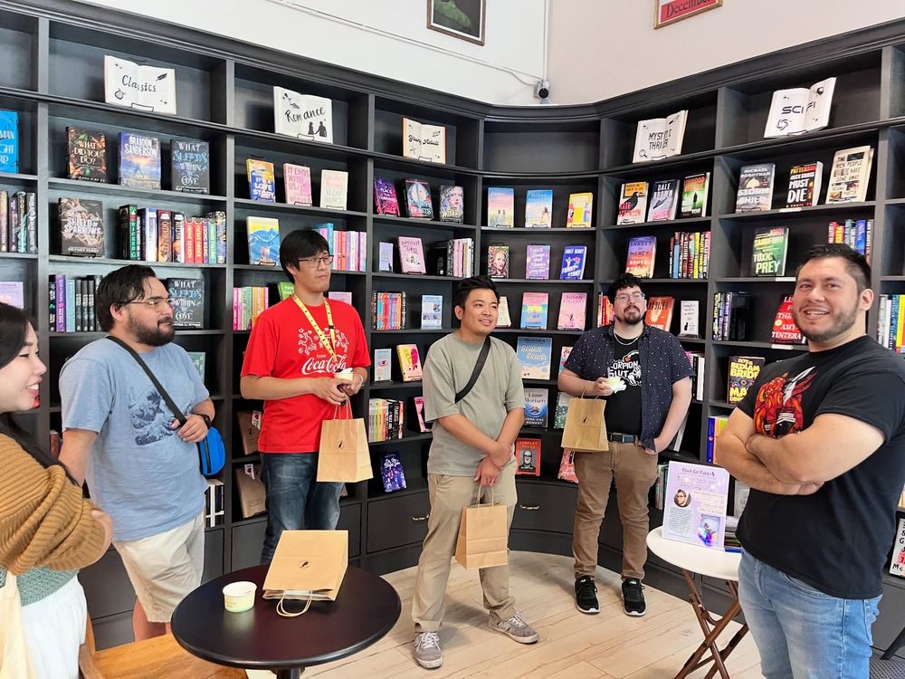 A group of readers in Black Cat Fables Bookstore with author Pedro Iniguez off to the side, smiling, arms crossed. The readers are all looking at him.