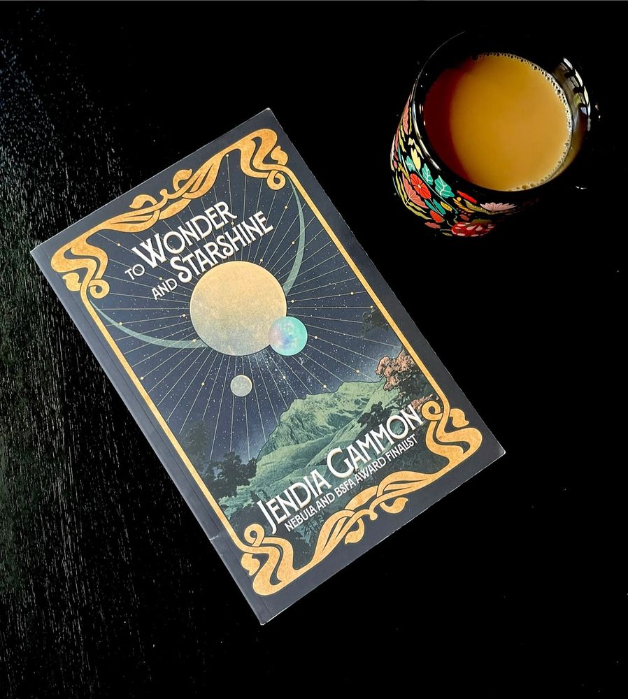 A paperback copy of TO WONDER AND STARSHINE by Jendia Gammon, a short story collection published by Trepidatio Publishing. It sits on a black table next to a black, floral, and insect design mug full of coffee.
