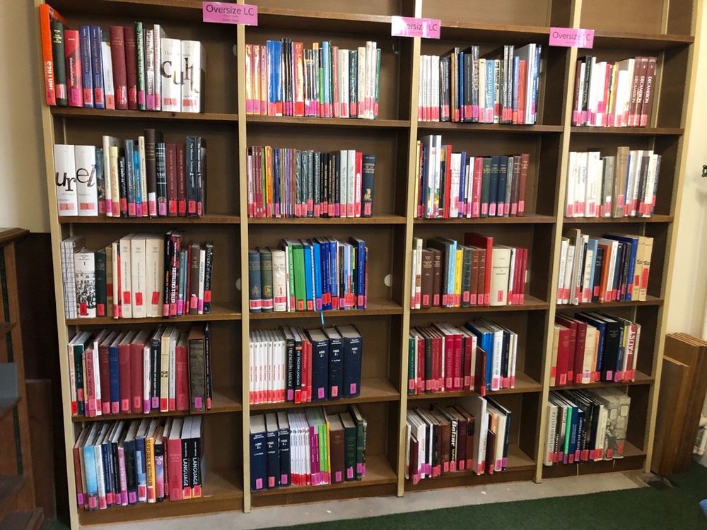 A picture showing the new location of the Teaching Collection Oversize books. The photograph shows 4 slim wooden bookcases lined up in a row. All of the shelves are full of books, and each book has a pink sticker on the spine.