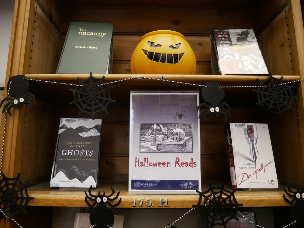 Two displays shelves of a Halloween book display. A poster in a plastic stand in the middle of the shelf reads 'Halloween Reads' in red gothic font, with an image of a linocut illustration of a skull resting on a book, next to a lit candle in a library. There is spider bunting along the shelves, and a plastic jack-o-lantern bucket on the top shelf. Four books can be seen, a green leatherbound book with the title 'the uncanny', a hardback with the title 'ghosts', a slavonic book with a fountain pen on the cover.