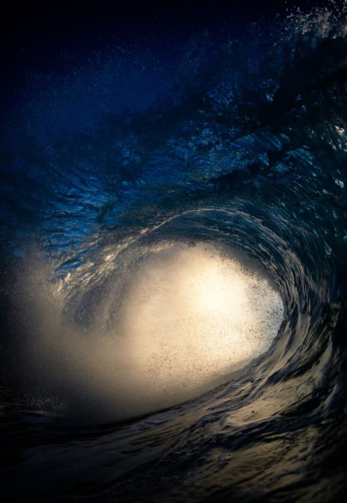 An dark ocean wave flares out over a reef as the sun lights up its vortex