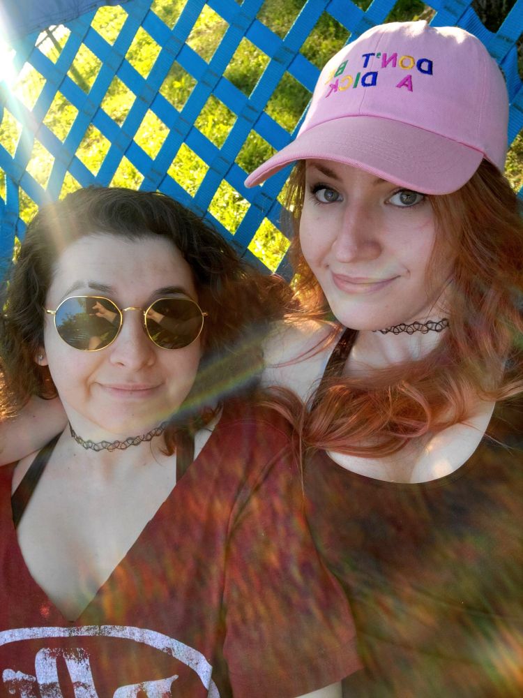 two girls smiling up at the camera while cuddling together in a hammock, one with curly dark hair and round sunglasses wearing a black choker and a v-neck dr. pepper t-shirt, the other with long wavy reddish hair under a pink baseball cap that says "don't be a dick" and wearing the same choker and a black athletic tank top