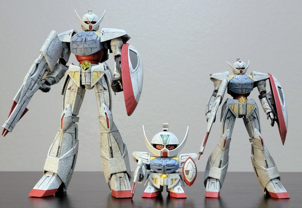 Three different gunpla models of WD-M01 ∀ Gundam (Turn A Gundam). They are all fully repainted in their standard colors. They are standing on a black table with a white wall in the background. The kits are the MG 1/100, SD, and HG 1/144.