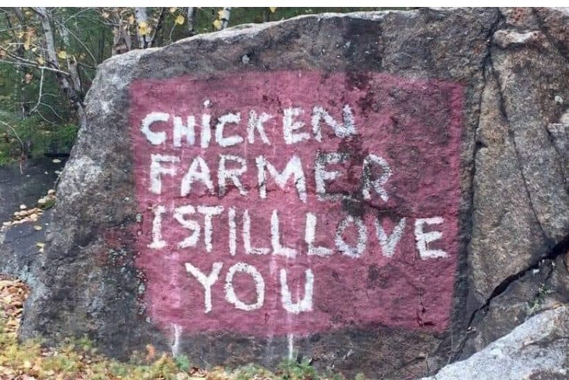 A boulder with a red painted square on it. In the square are white painted letters that say “CHICKEN FARMER I STILL LOVE YOU”