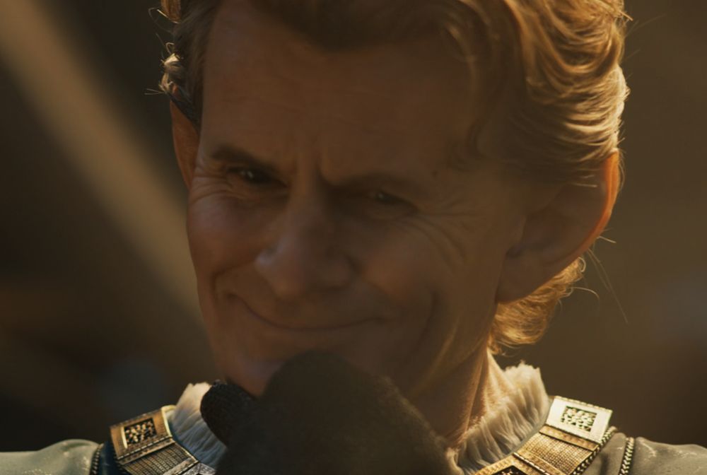 Uma foto de Celebrimbor na série Os Anéis de Poder. Ele está com o queixo apoiado em uma das mãos, com um grande sorriso no rosto, enquanto olha para alguma coisa que não aparece na imagem, como se orgulhoso.