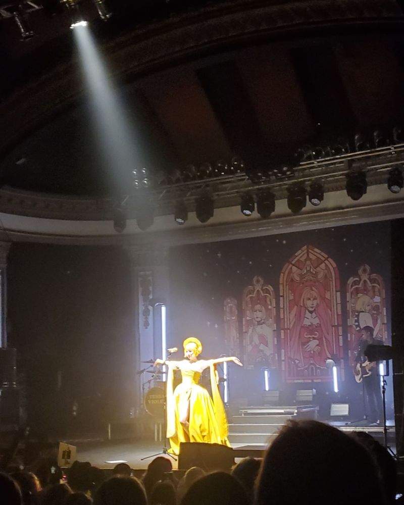 Musician and songwriter Tessa Violet is pictured onstage at the Newport Music Hall in Columbus Ohio. She is wearing a bright yellow vinyl gown and halo inspired headdress. Her arms are outstretched and she is lit by a single spotlight from above.