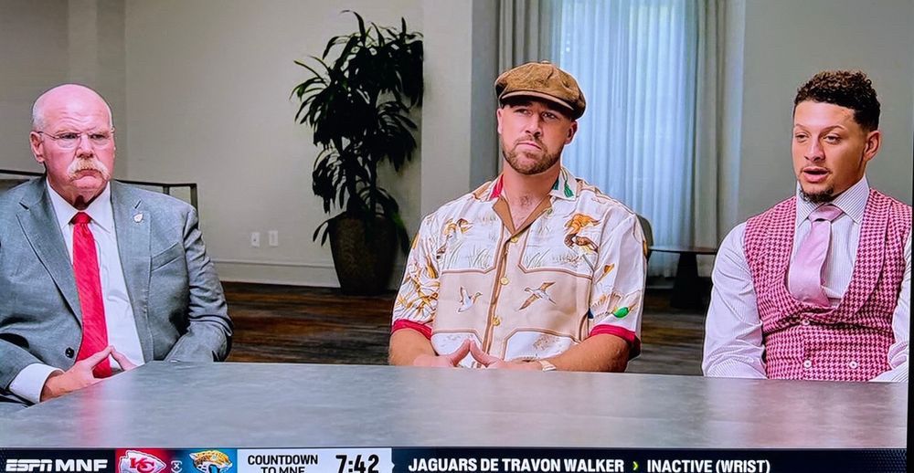 Andy Reid in grey suit, Travis Kelce in bird print hawaiian shirt and cap, Patrick Mahomes in fancy pink shirt-vest-tie