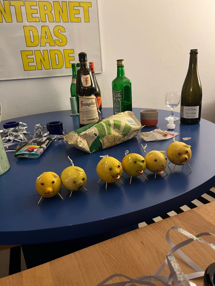 sechs zitronenschweine stehen auf einem blauen tisch umgeben von verschiedenen flaschen, drehzeug und chips. im hintergrund ein plakat mit gelber schrift auf weißem grund, es sind die worte „internet das ende“ zu erkennen 
