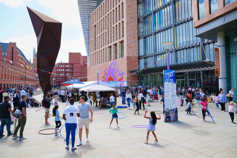 A large group of adults and children are engaging in various activities, including some using hula hoops, outside a large building in the sun.