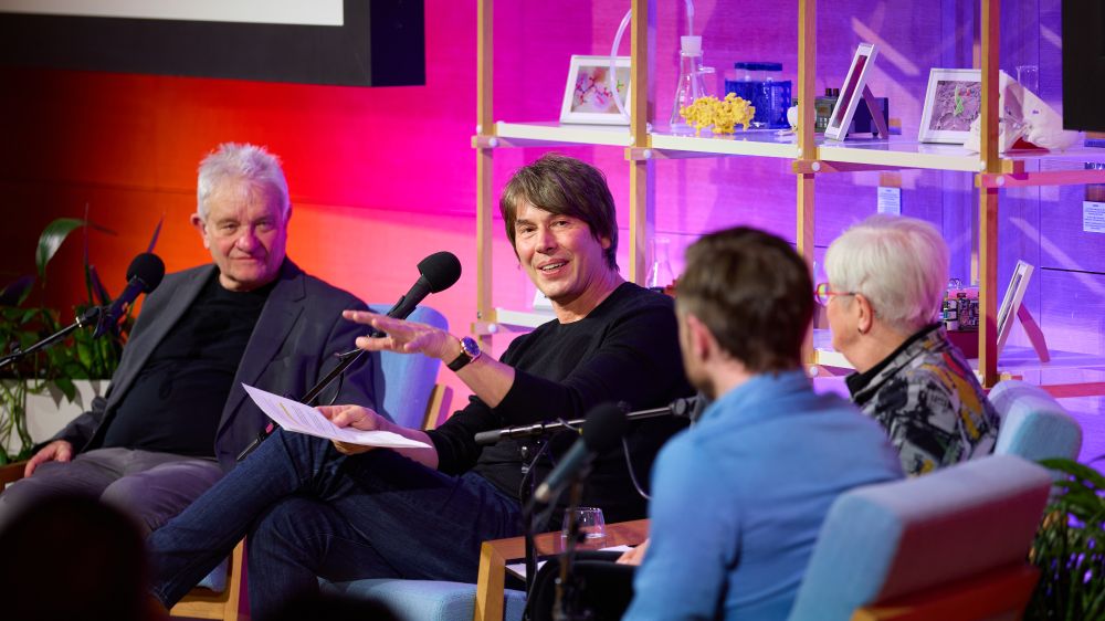 Brian Cox sits in a well-lit chair in front of a bookshelf. He has a microphone in front of him, and three other guests are sitting around him, also with microphones in front of them.