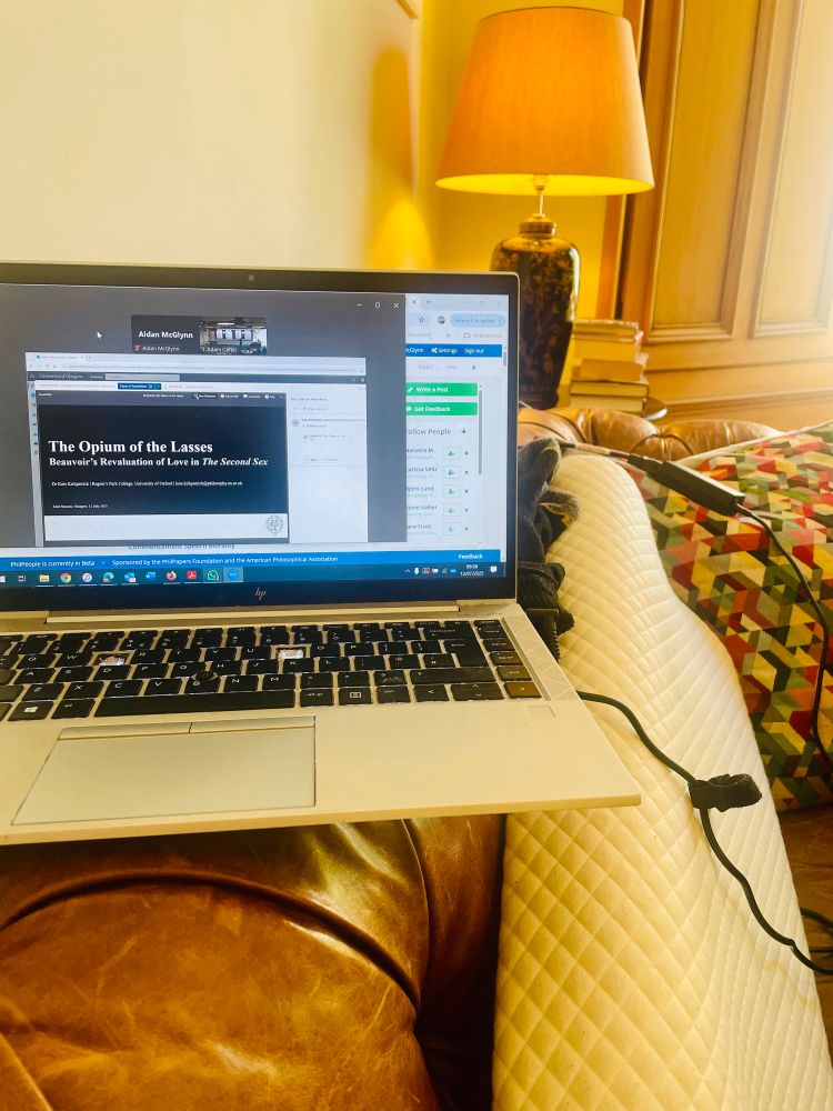 A laptop showing zoom with a slide on Beauvoir in love in the Second Sex, sitting on the back of a brown leather couch