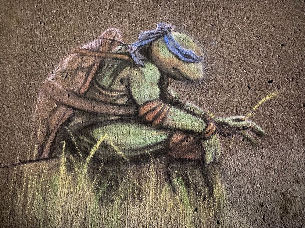 A chalk drawing of Leonardo (the ninja turtle) sitting low, possibly on a log in some dry grass, head down in concentration or contemplation. He holds a piece of dry grass in his left hand, his right is resting on his knee.