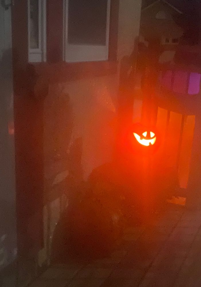 A lopsided-smiling jack-o-lantern seen through a fogged up window. Still lit from within by a candle’s glow. 