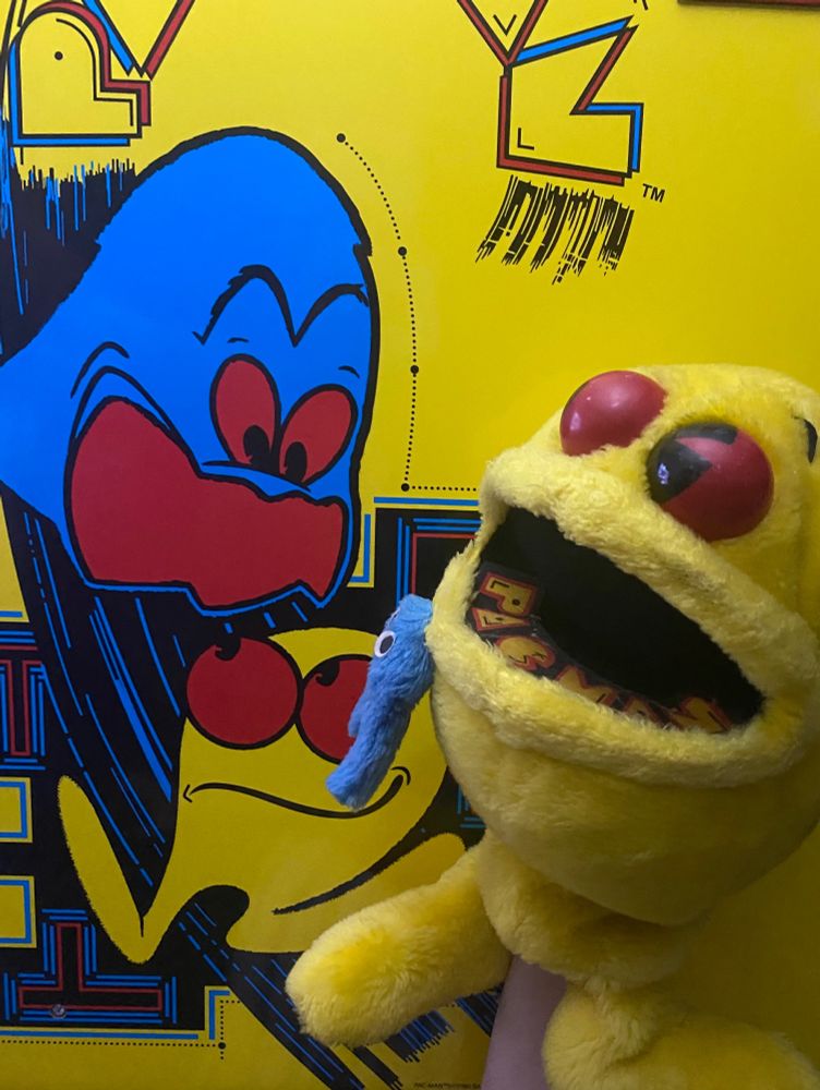 A vintage 80’s PAC-Man puppet in front of a 1-Up PAC-Man arcade. The puppet has legs and large red eyes, like the illustration on the cabinet. 