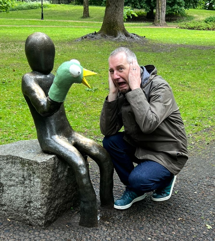 A photo of me next to the statue Ansiktsløse Mann by Evald Møller Johansen. The statue is a faceless figure holding a hand puppet of a bird. 