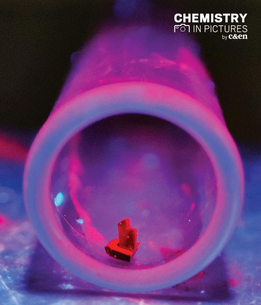 A photograph of a red, vaguely boat-shaped crystal in a test tube under ultraviolet light. 