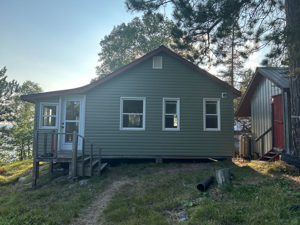 This is Ward point cabin. It is Moss green with three white windows and a white door made of glass to the right is an outhouse.