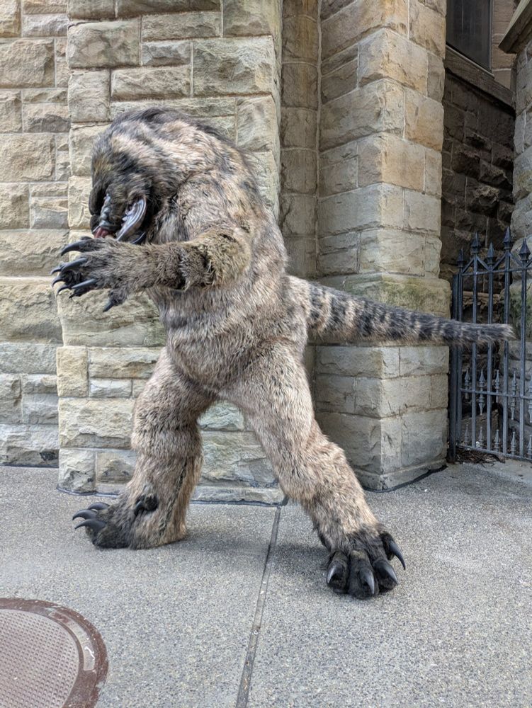 View from the side of a snarling monster with big teeth, big claws, and buglike mandibles on its face striking a threatening pose in front of a stone building