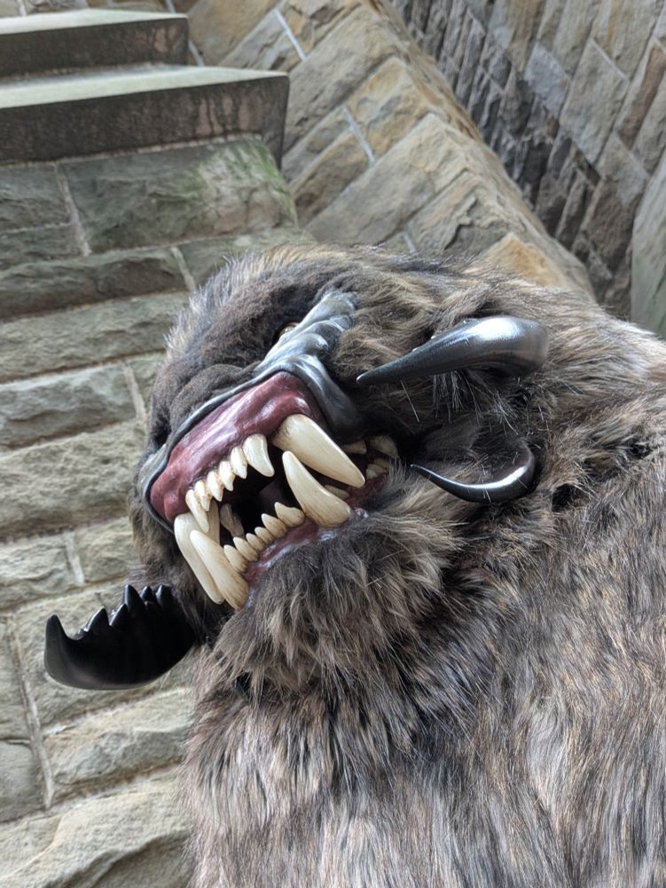 View from below of a snarling monster with big teeth and buglike mandibles on its face, in front of a stone building