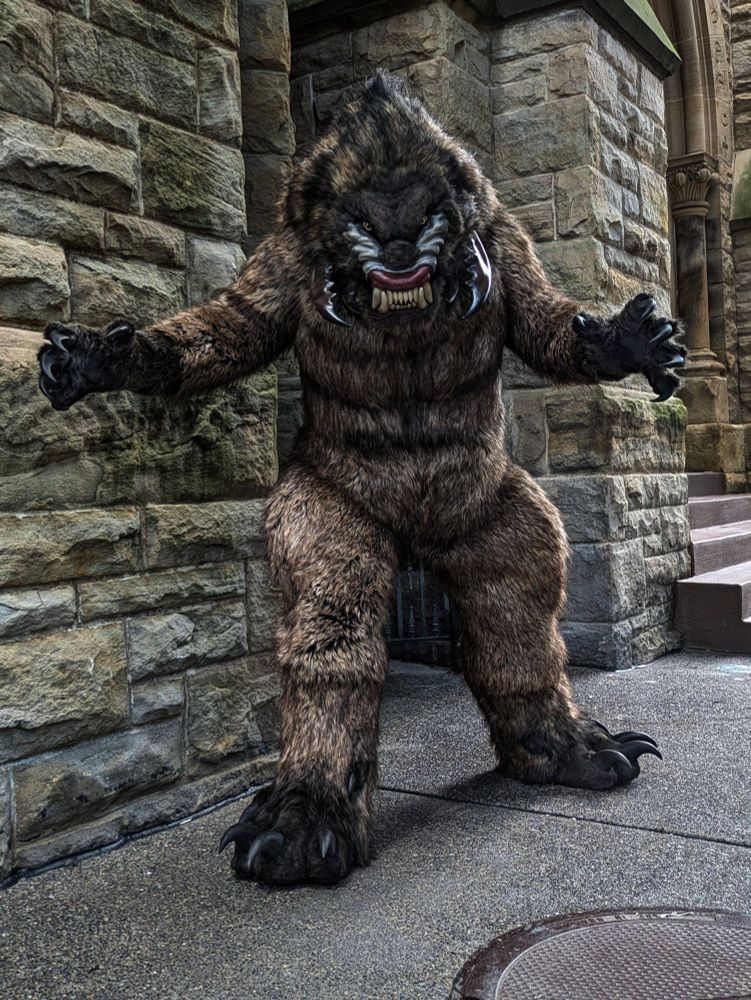 A snarling monster with big teeth, big claws, and buglike mandibles on its face strikes a threatening pose in front of a stone building