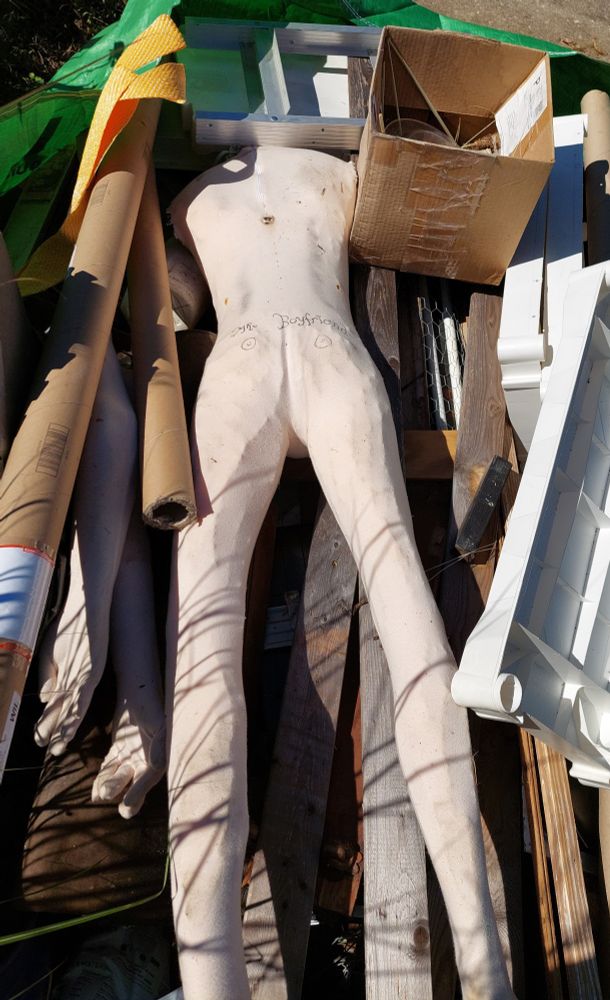 A bagster full of junk. Among the junk is a dingy, rough foam mannequin body with a cloth skin, lying face-down. Drawn on its butt in sharpie are a pair of nipples and the words "Mr. Boyfriend" in a swoopy style with the dots replaced with little hearts.