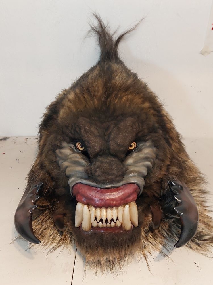 A brown-furred monster head with a mandrill-like snarl, intense yellow eyes, and crab-claw-like facial extensions. Front view.