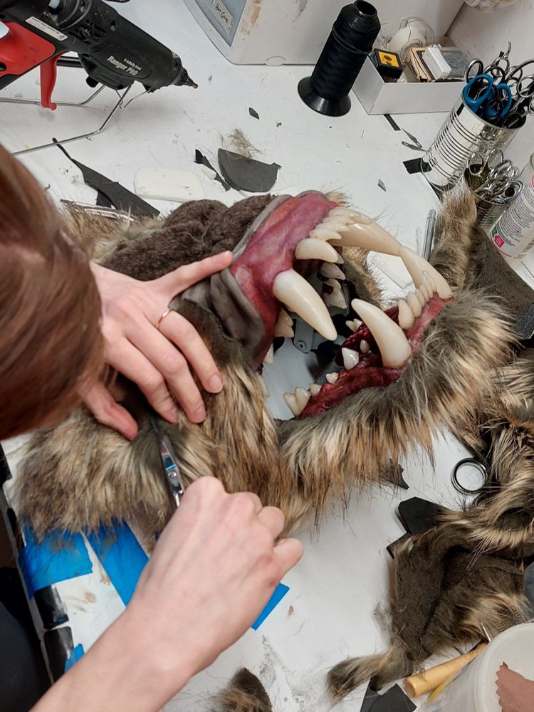 A monster mask lies face-up on a cluttered table, being actively worked on by Shanzi wielding a pair of scissors. Its huge teeth and retracted lips, framed by brownish fur, give an impression of a snarling mandrill.
