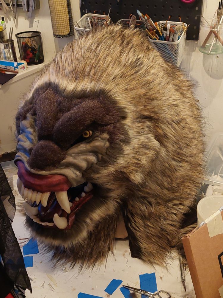 A brown-furred monster mask on a cluttered work table. It is very large. Its snarling face with huge bared teeth and hairless cheeks is reminiscent of a mandrill. 