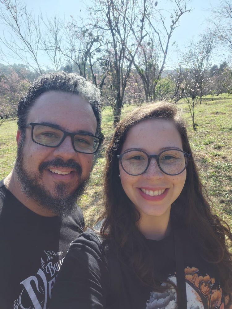 Selfie de um casal, homem branco de óculos e barba e mulher branca de óculos. Com árvores ao fundo, grama e céu azul. 