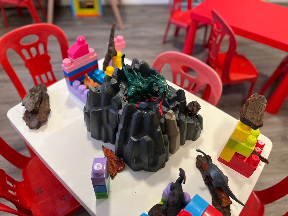 Large colourful blocks, bark, and plastic dinosaurs surround a low toy mountain with a dragon on top, breathing fire downward.  They are all on a white rectangular table set with four chairs