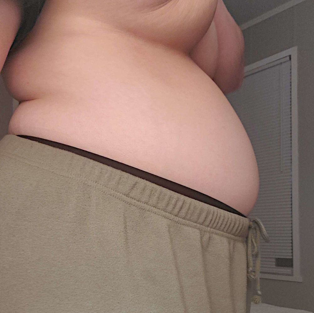 photo of a taut belly in grey green lounge pants, the band of panties peaking out from under the pants. the persons boobs are being pulled up from their chest to exaggerate the curve of the belly