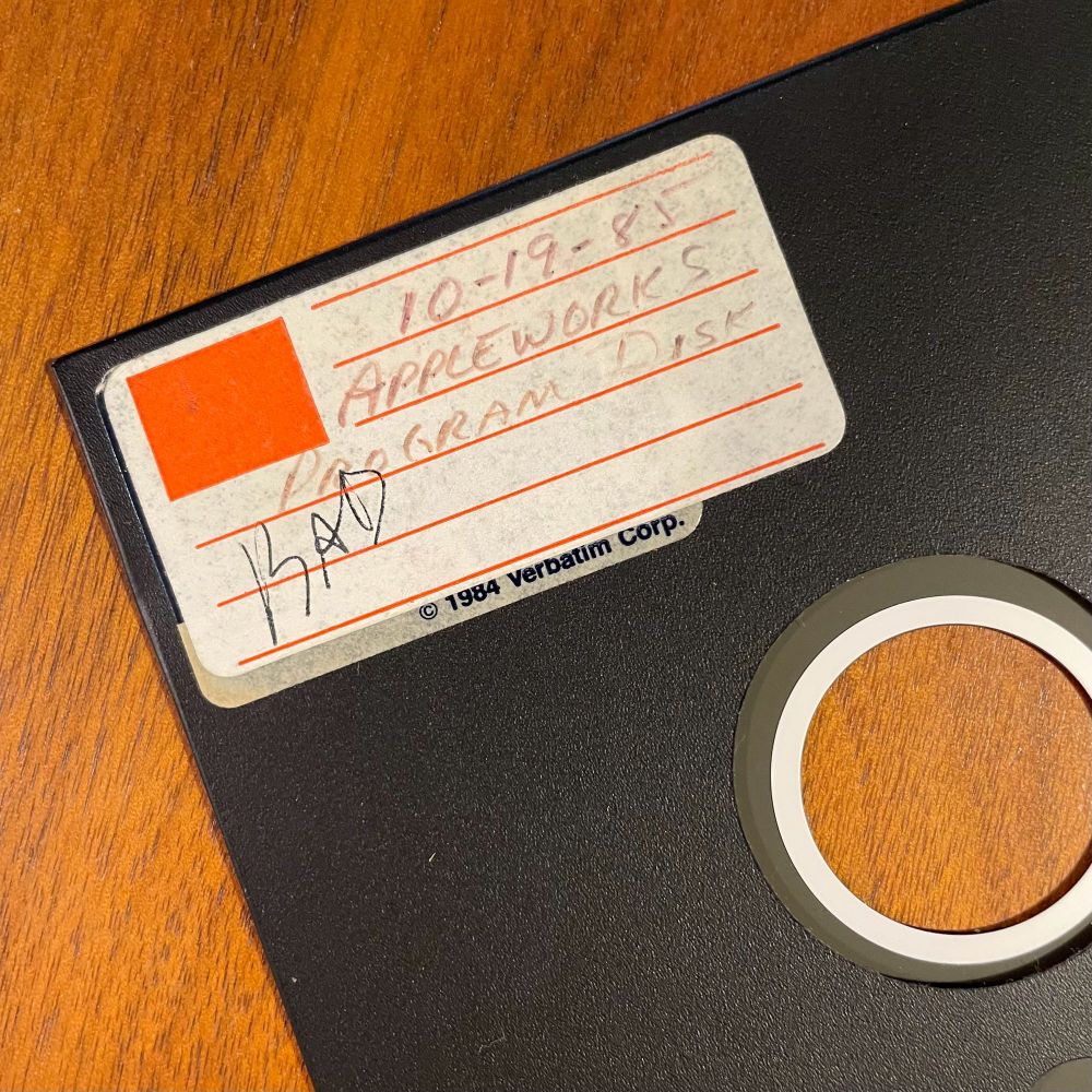 A 5.25” floppy diskette on a wood desk with a label that says 
“10-19-85
APPLEWORKS PROGRAM DISK”, and also “BAD” in a different color ink