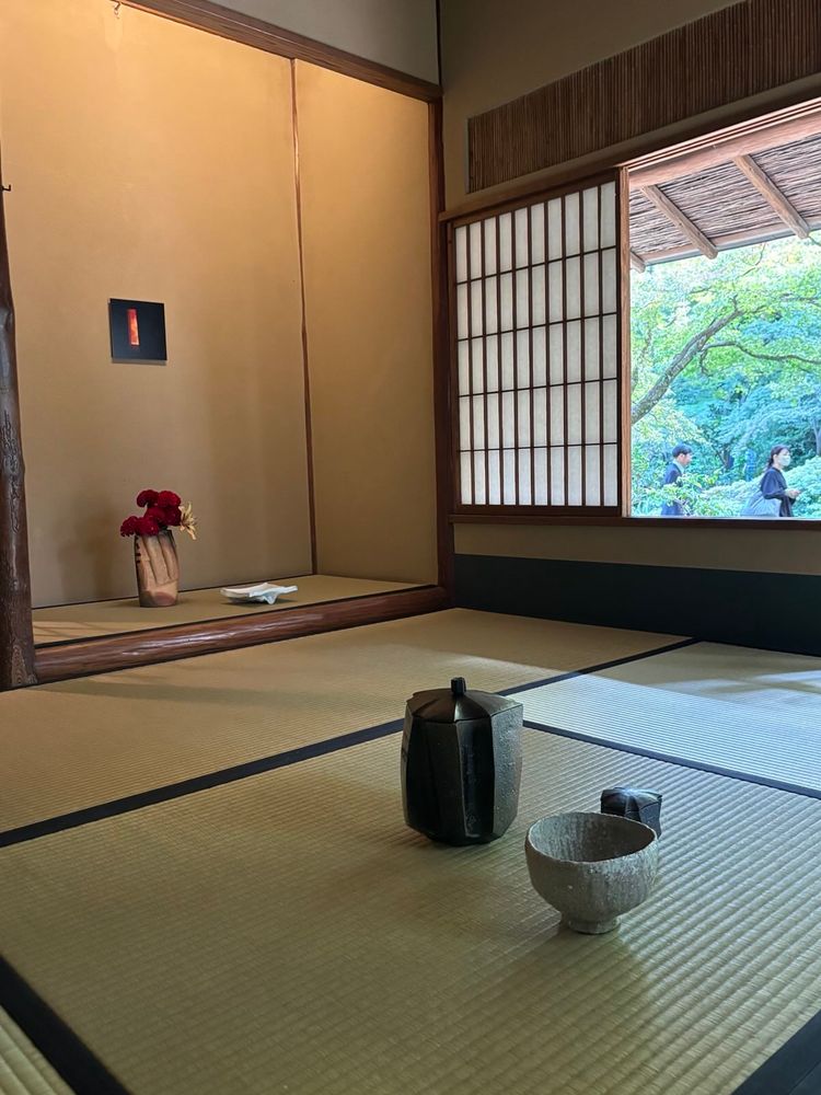 Schlichter Teeraum mit Ausblick in den Garten. Auf dem Boden stehen eine Macha-Schale, ein kleines Gefäß (für den Tee vermutlich) und eine Wasserkanne. In einer Nische im Hintergrund stehen Blumen in einer Vase und ein Bild hängt an der Wand.