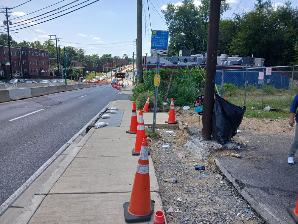 In Silver Spring, Maryland, view west along north side of Piney Branch Road east of University Boulevard. In foreground, a bus stop sign. No shelter, seating, or receptacles are visible.