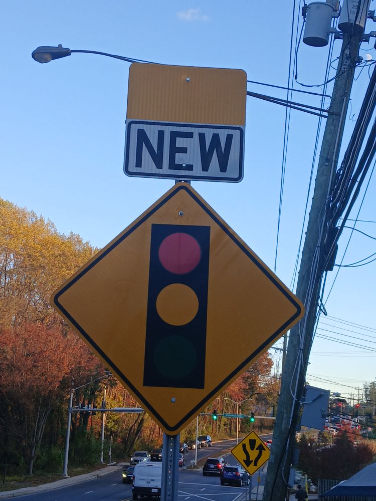 In Adelphi, Maryland, view east on Adelphi Road west of Edwards Way. In foreground, a sign pole with, from top, a yellow rectangle, a black-on-white sign with the word "new," and an orange diamond-shaped sign with a traffic signal icon.