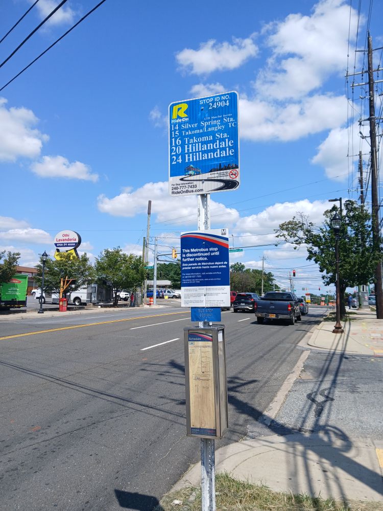 In Silver Spring, Maryland, on Piney Branch Road near Flower Avenue (south side), a sign pole with (from top) a Ride On bus stop sign, a Metrobus stop closure notice, a Metrobus Braille placard, and a Metrobus J4 timetable.