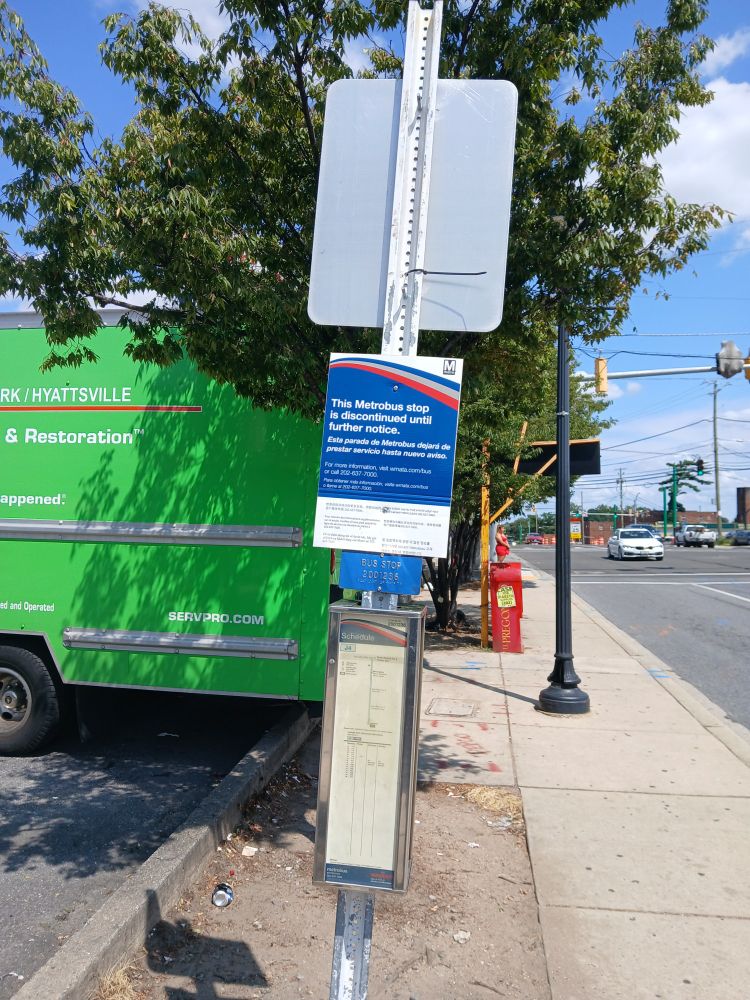 In Silver Spring, Maryland, on Piney Branch Road near Flower Avenue (north side), a sign pole with (from top) the back of a Ride On bus stop sign, a Metrobus stop closure notice, a Metrobus Braille placard, and a Metrobus J4 timetable.
