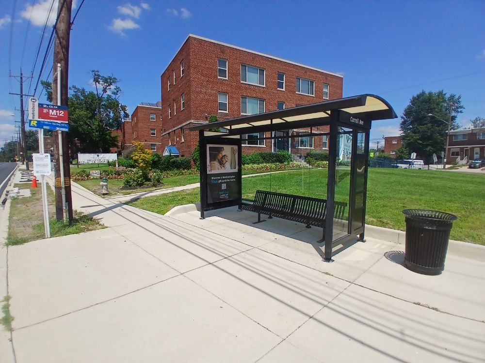 In Silver Spring, Maryland, view west along north side of University Boulevard west of Carroll Avenue. In foreground, a bus stop shelter. A trash receptacle is visible; no recycling receptacle is visible.