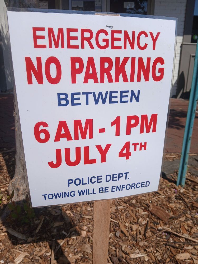 On a wooden stake, a roadside sign withthe text "Emergency parking between 6AM - 1PM July 4th. Police Dept. Towing will be enforced."