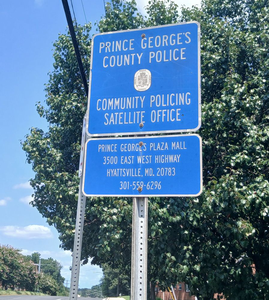 In Prince George's County, Maryland, on Riggs Road, stacked roadside blue-on-white signs including the text "Community Policing Satellite Office" and "3500 East West Highway."
