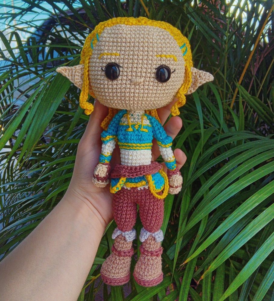 Amigurumi de Zelda de The Legend Of Zelda: Breath Of The Wild sendo segurada por uma mão com folhas de coqueiro ao fundo.