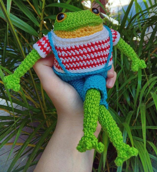 Foto de um amigurumi de um sapo verde usando uma camisa listrada branco e vermelha e uma jardineira azul 