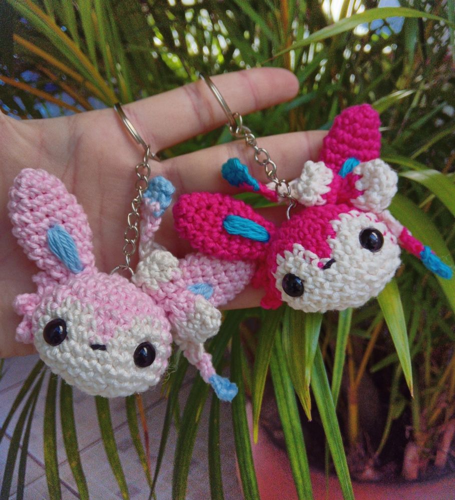 Foto de dois chaveiros de Amigurumi de cabeças de Sylveon, a evolução fada do Evee. Na esquerda temos a cabeça com cores pastéis e na direita temos a cabeça com cores mais saturadas 