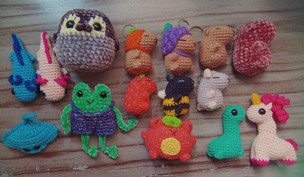 Foto com 15 amigurumis. Temos dois axolotes e um é azul e o outro rosa, um pinguim, capivara com chapéu de laranja, uva, sem chapéu, pelúcia, toda morango, abelha e rato, arraia, sapo com jardineira, sapo morango, dinossauro e unicórnio 